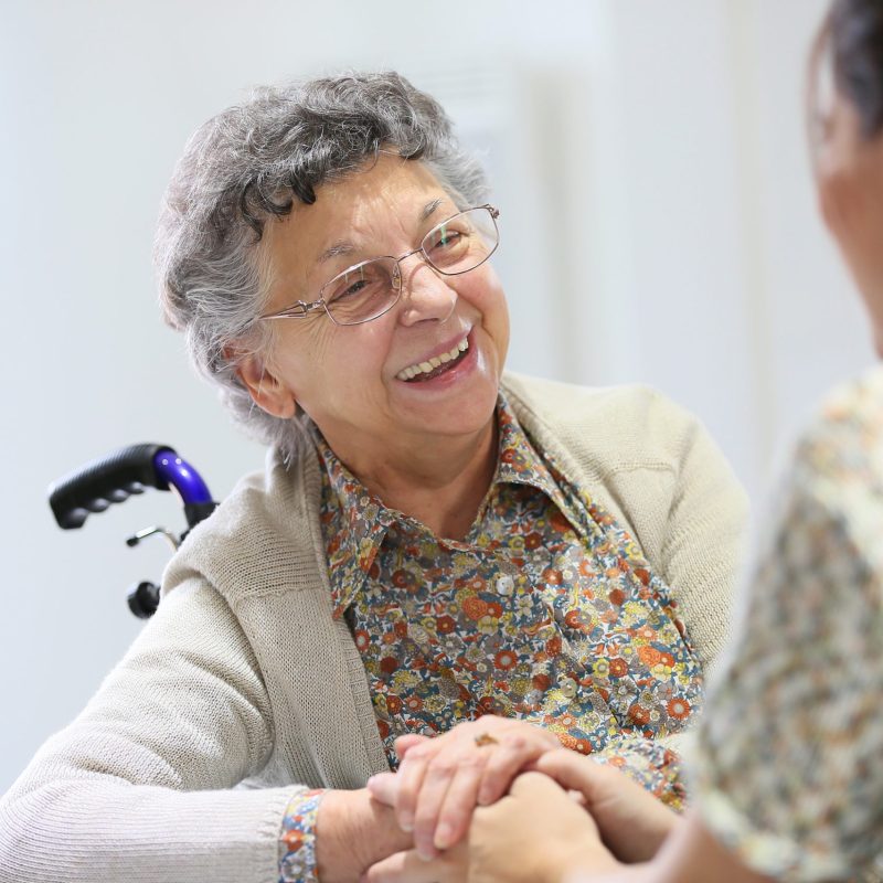An older woman sitting in a wheelchair and happily holding the hand of her caretaker